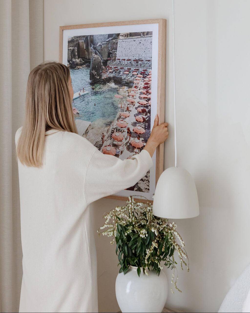 Woman hanging a framed coastal Italy wall art print by The Print Emporium on a wall with a vase and plant in the foreground.